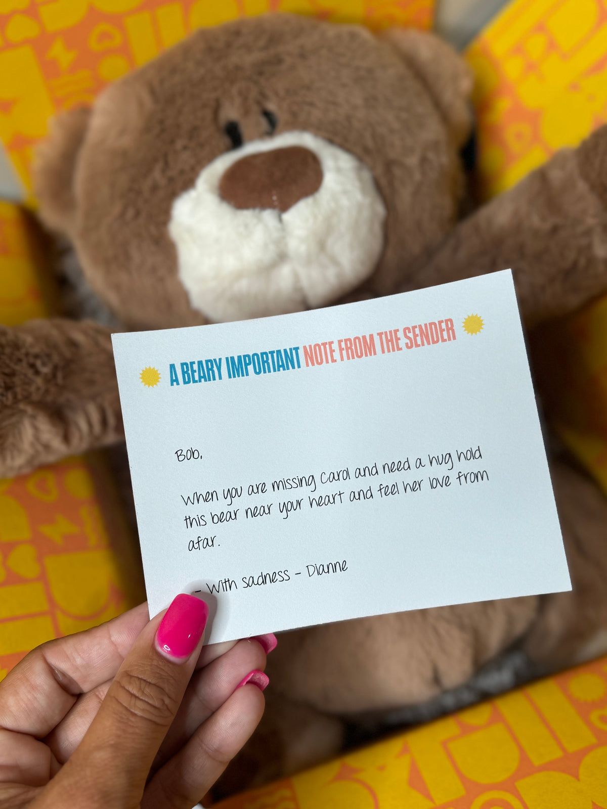 A hand with pink nail polish holds a white note card in front of a brown teddy bear, with a yellow patterned background.